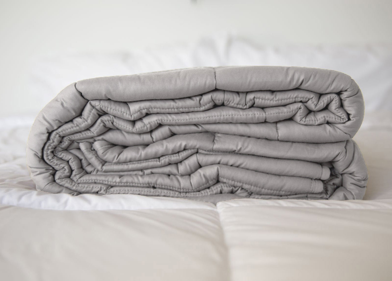 A neatly folded Pillowtex Weighted Blanket in gray rests atop a white, quilted bedspread in a softly lit room.